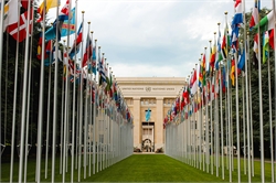 [ai] A pathway lined with flags of various countries leading to the United Nations building in Geneva, surrounded by green grass and trees under a cloudy sky.